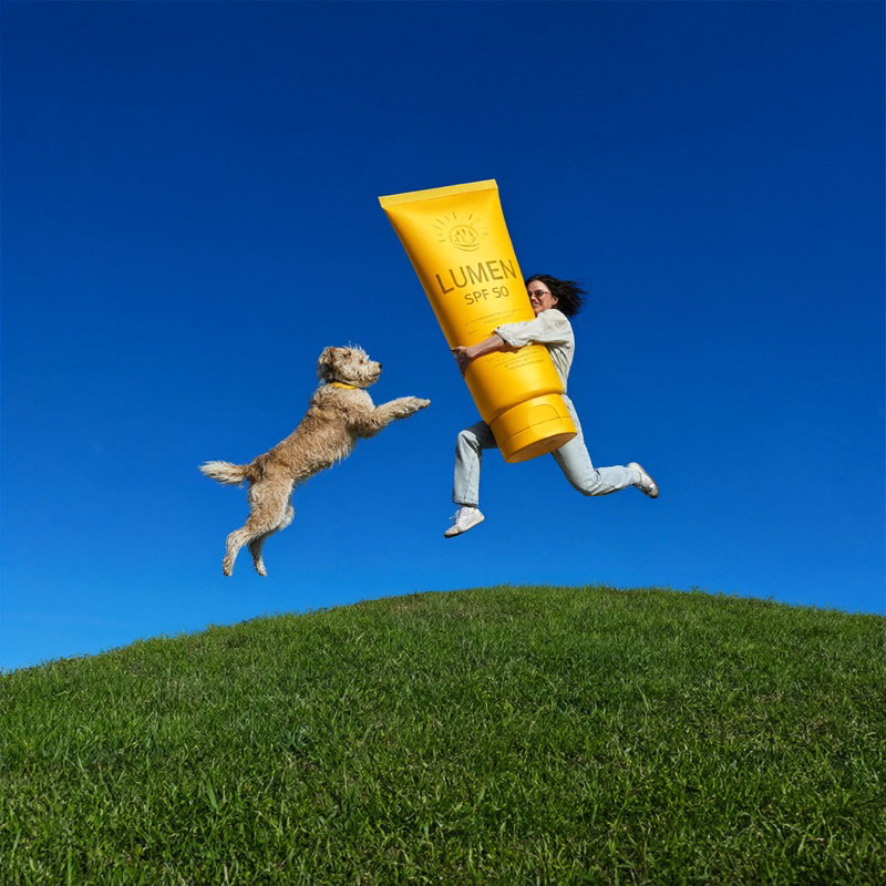 A LUMEN sunscreen ad: a person and a dog leap after a giant yellow tube