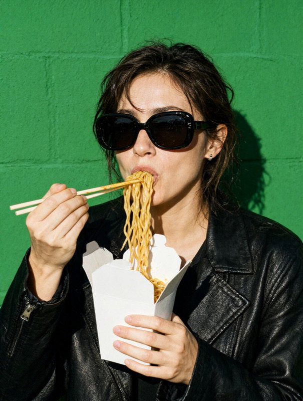 A woman in sunglasses and a leather jacket eats noodles in front of a green wall
