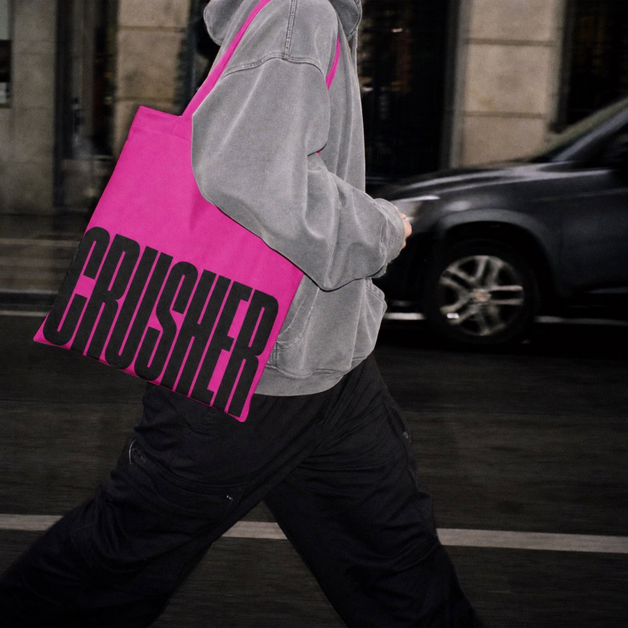 A person in a gray hoodie carrying a pink CRUSHER tote bag across a street